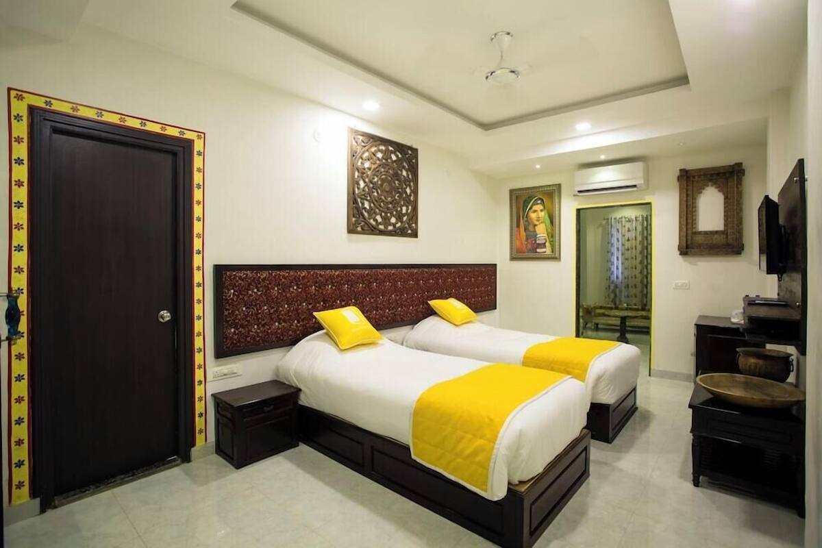 A hotel room with two beds, a dark wooden door, a decorative wall piece, a mirror, and a small table.