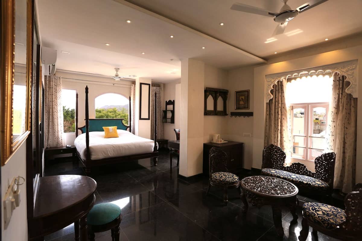 A spacious bedroom with a four-poster bed, a sitting area with ornate chairs, and a view of greenery outside.