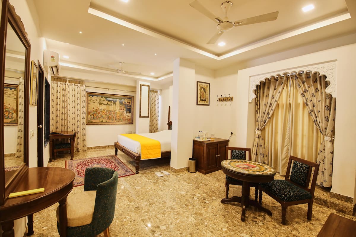 A hotel room with a bed, a dining table, a chair, a small table with a decorative item, a mirror, and a ceiling fan.