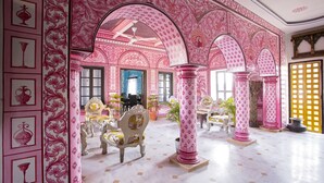 A room with pink floral wallpaper, white marble flooring, and ornate columns. There are several chairs and a table with a vase.