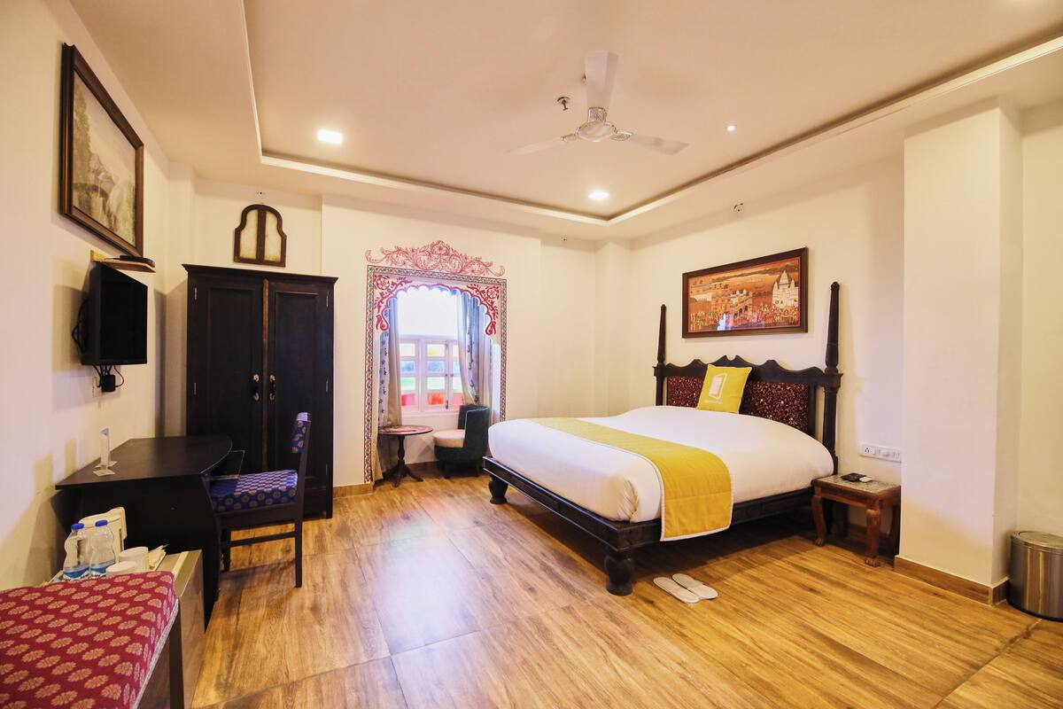 A hotel room with a wooden floor, a bed with a yellow blanket, a desk with a chair, a wardrobe, a small table, and a framed picture on the wall.