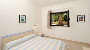 Un dormitorio con una cama, una mesita de noche, un ventanal con vista a los árboles y dos cuadros en la pared.