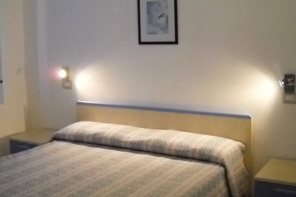 Siististi pedattu sänky, jossa on kuviollinen päiväpeite, kaksi yöpöytävalaisinta ja seinällä oleva kehystetty kuva.