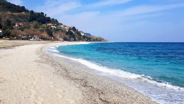 A beach with pebbly shore, clear blue water, and houses on a hillside in the background. - Katerina Fotopoulos Homes & Apartments (Zagora-Mouresi)