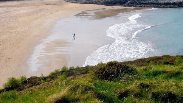 A beach with two people walking along the shore, surrounded by grassy hills and a calm sea. - Corcreggan Mill (Dunfanaghy)