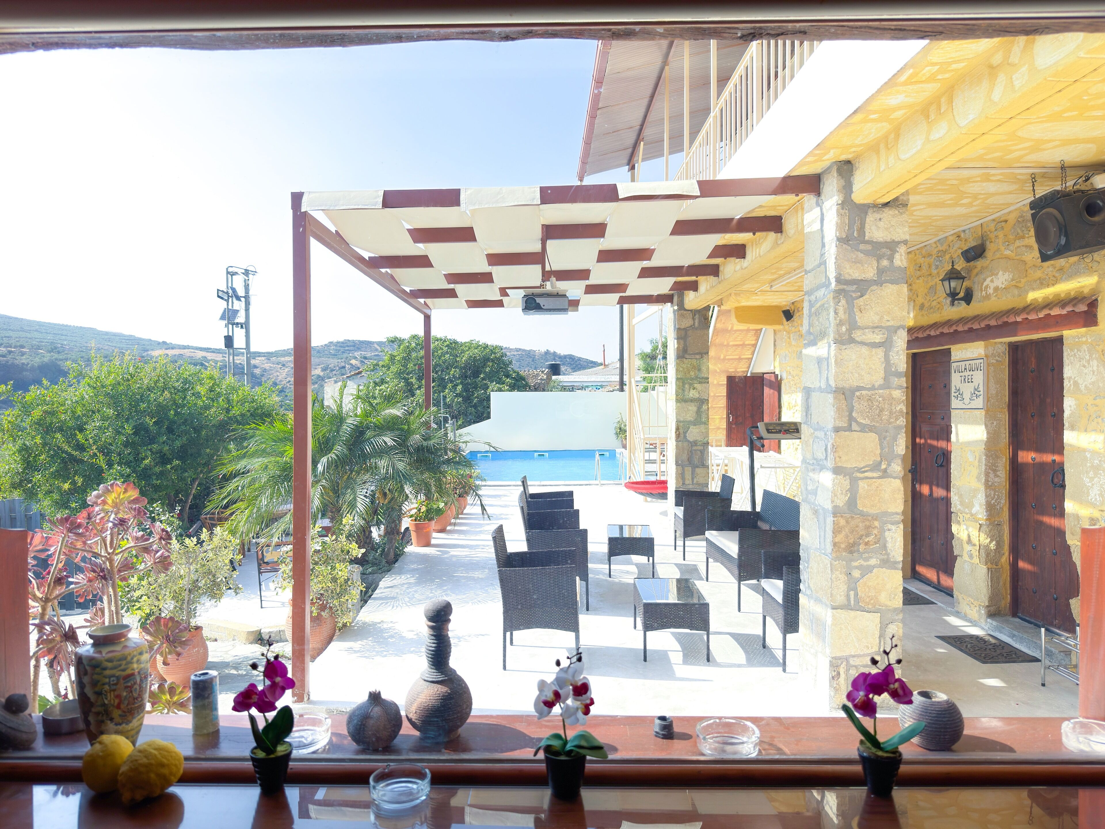 A terrace with outdoor seating, a view of the sea, and a stone building with a sign reading "Villa Olive Tree".