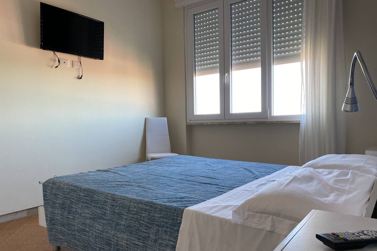 Ein Schlafzimmer mit Bett, einem an der Wand montierten Fernseher, einem Fenster mit Jalousien und einem Nachttisch mit Fernbedienung.