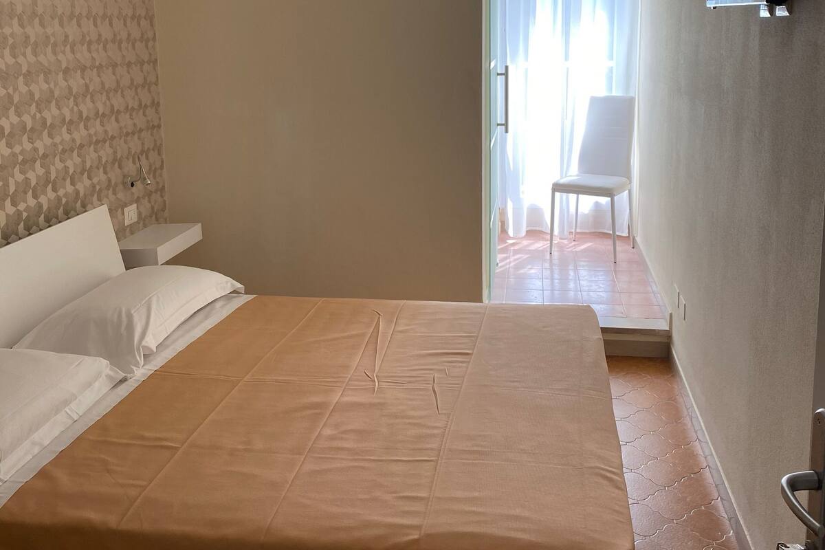 Ein Hotelzimmer mit Bett, Wandfernseher und Blick ins Freie durch ein mit Vorhängen versehenes Fenster.