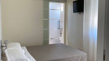 Un dormitorio con cama, un banco, un televisor y un baño visible a través de una puerta de cristal.