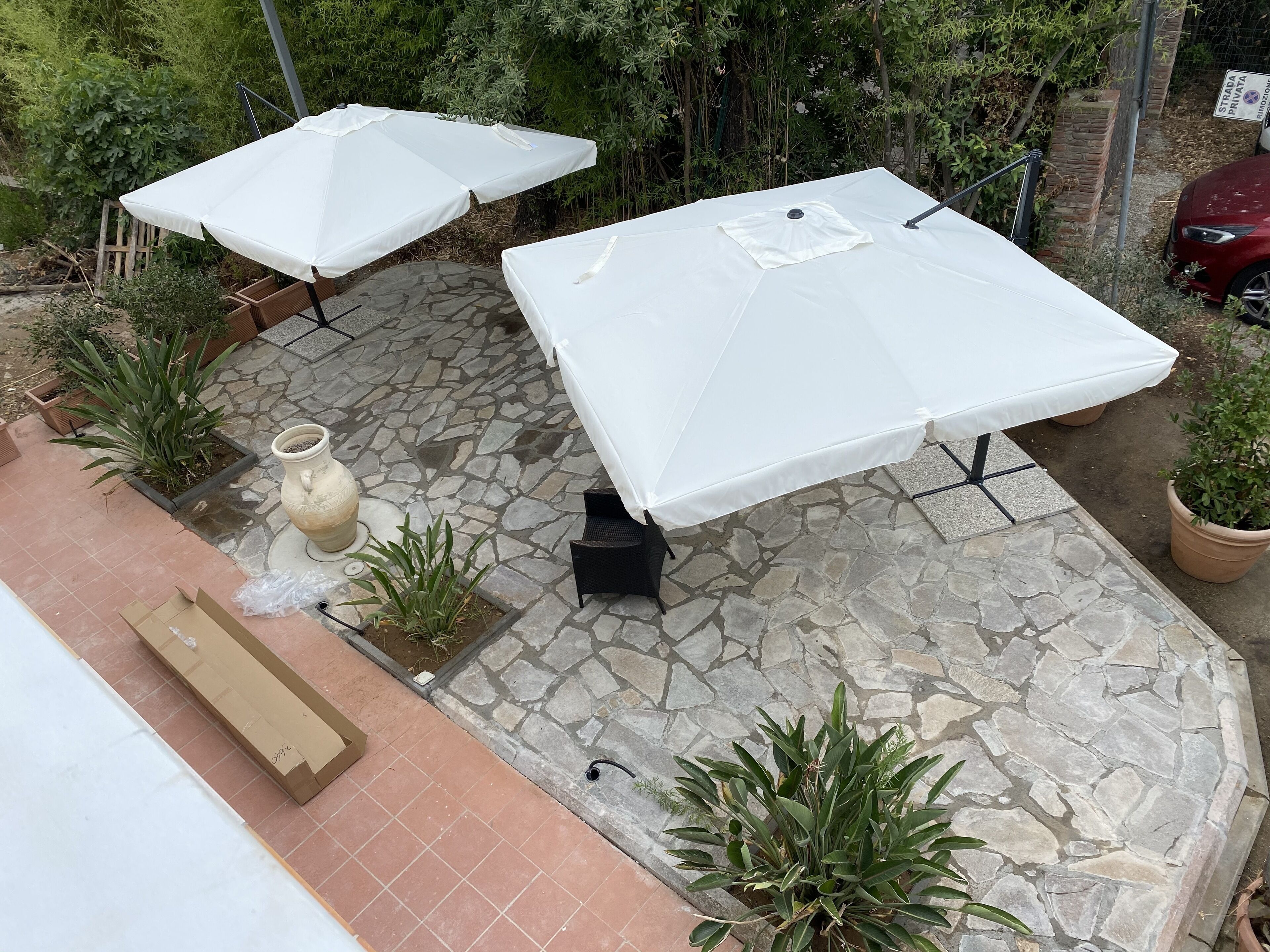 Un patio al aire libre con suelo de piedra, dos sombrillas blancas, una silla negra y plantas en macetas.