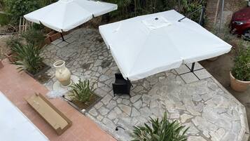 Un patio al aire libre con suelo de piedra, dos sombrillas blancas, una silla negra y plantas en macetas.