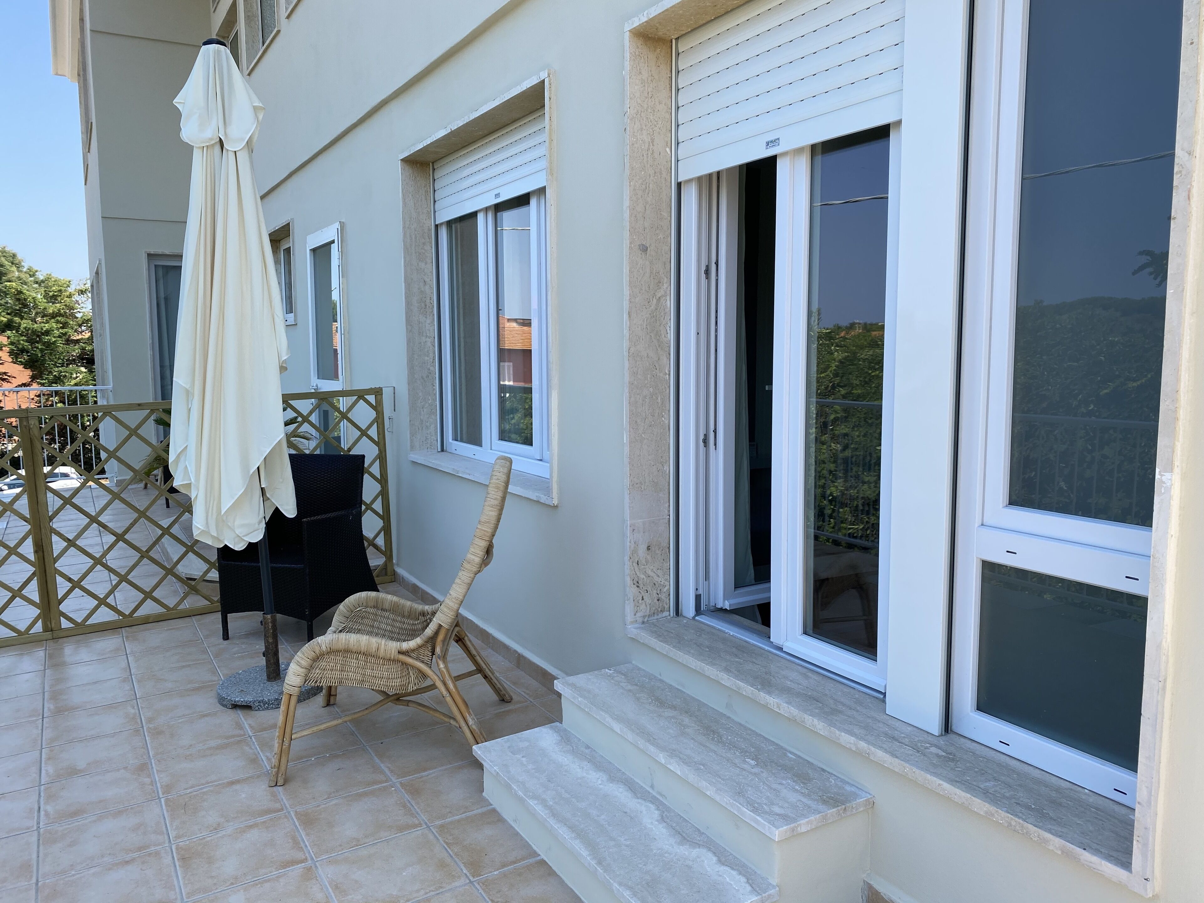 A balcony with a wicker chair, a white umbrella, and a view of the street.