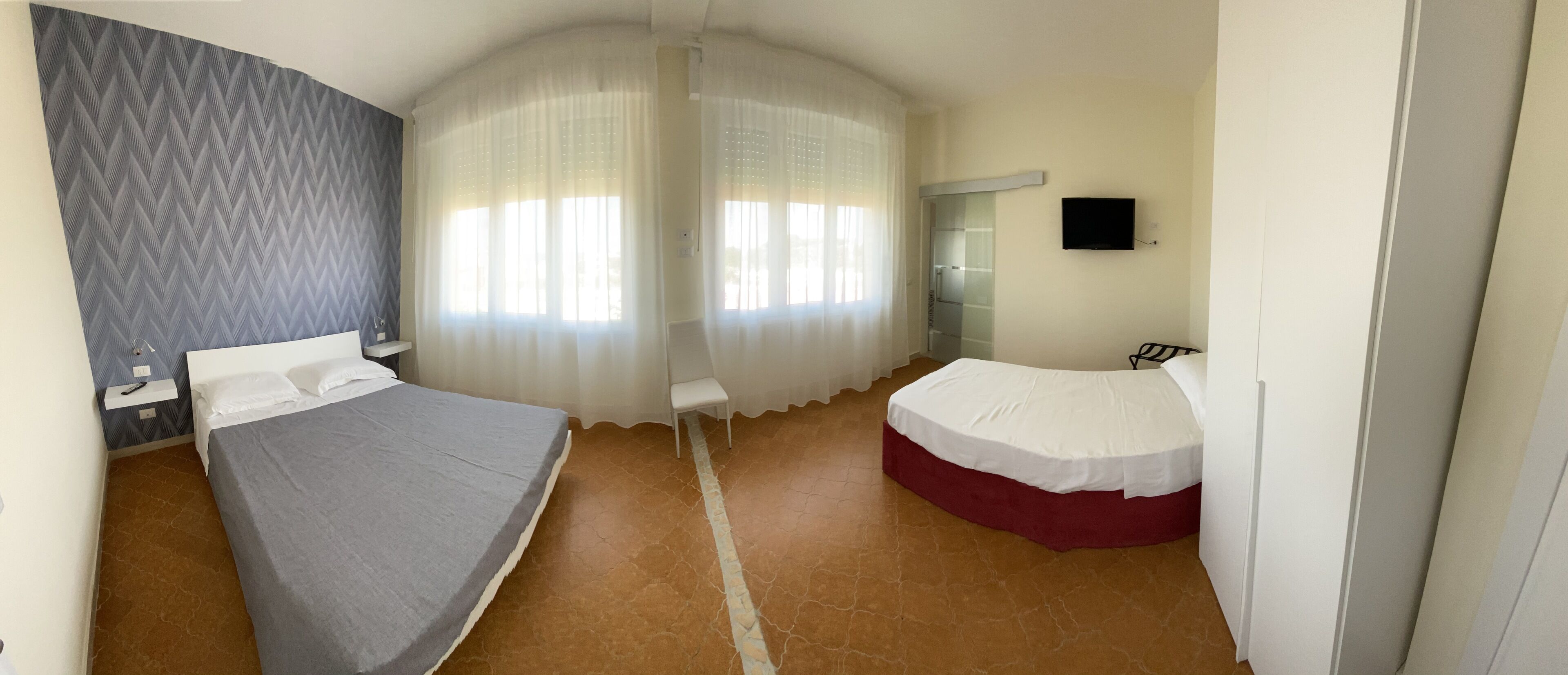 Ein Schlafzimmer mit zwei Betten, einem Spiegel, einem Fernseher und einem Fenster mit durchsichtigen Vorhängen.