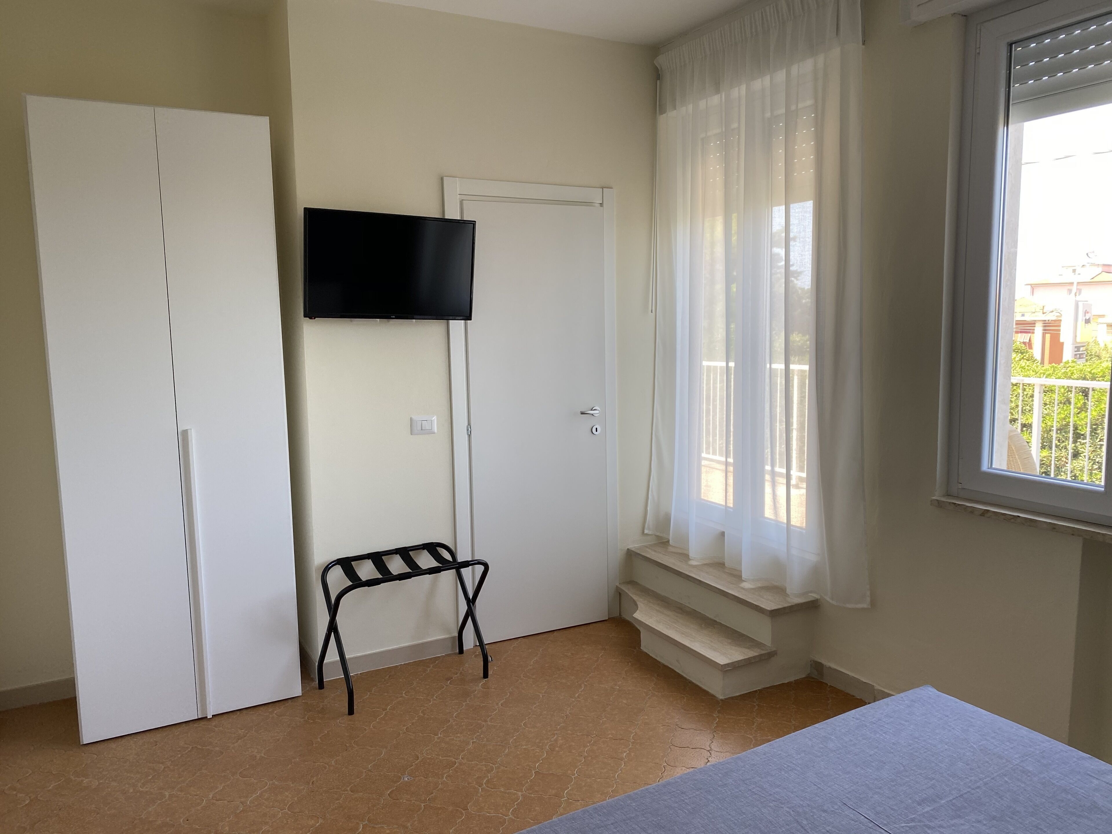 A room with a bed, a TV mounted on the wall, a window with sheer curtains, and a small table with a chair.