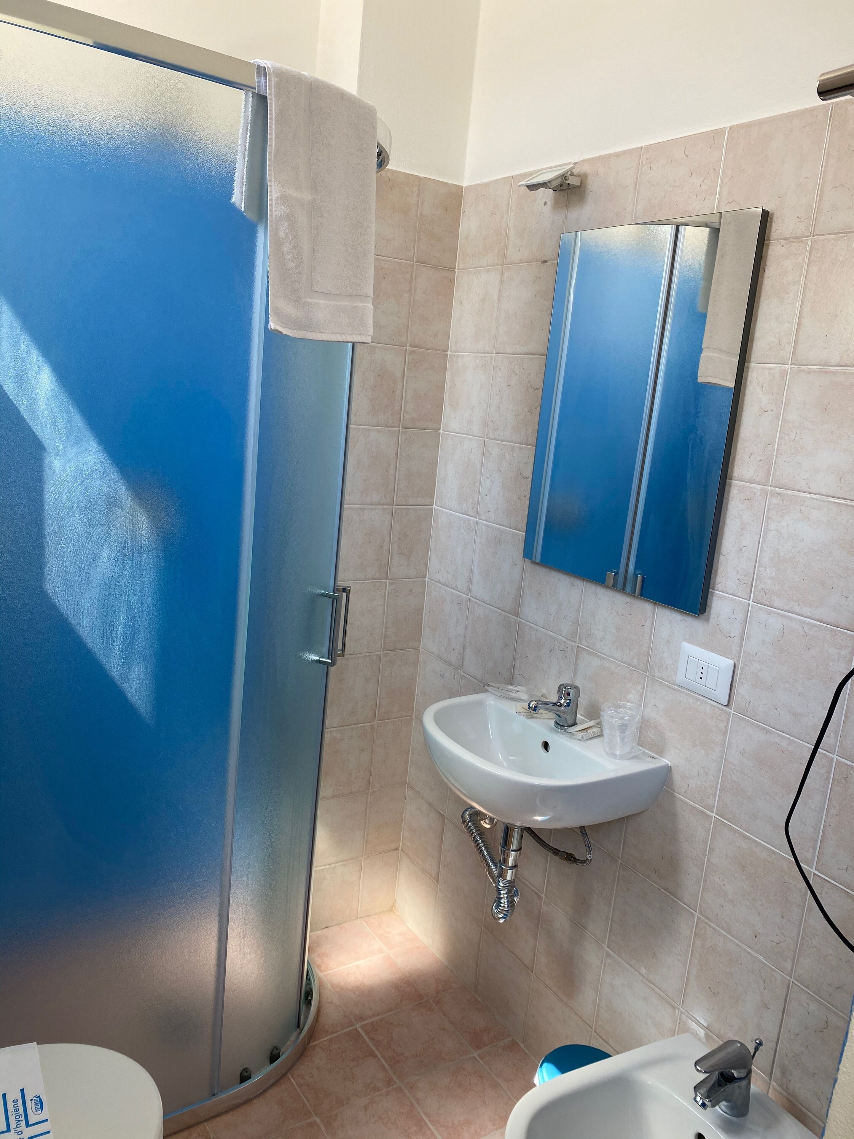Een badkamer met een blauwe douchecabine, een wastafel met spiegel erboven en beige betegelde wanden.