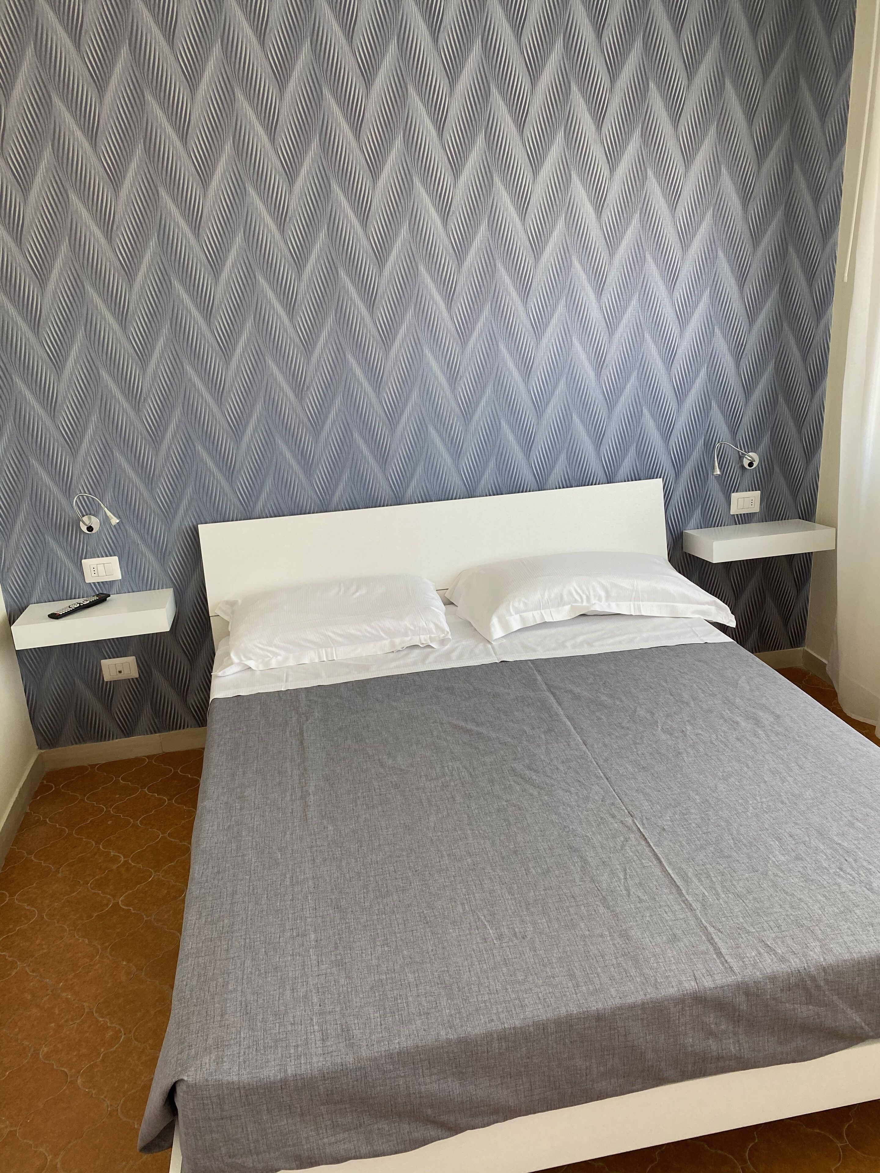 Ein ordentlich bezogenes Bett mit weißen Leinen, einer grauen Tagesdecke und einer gemusterten Tapete. Es gibt zwei Nachttische mit Lampen und eine Steckdose an der Wand.