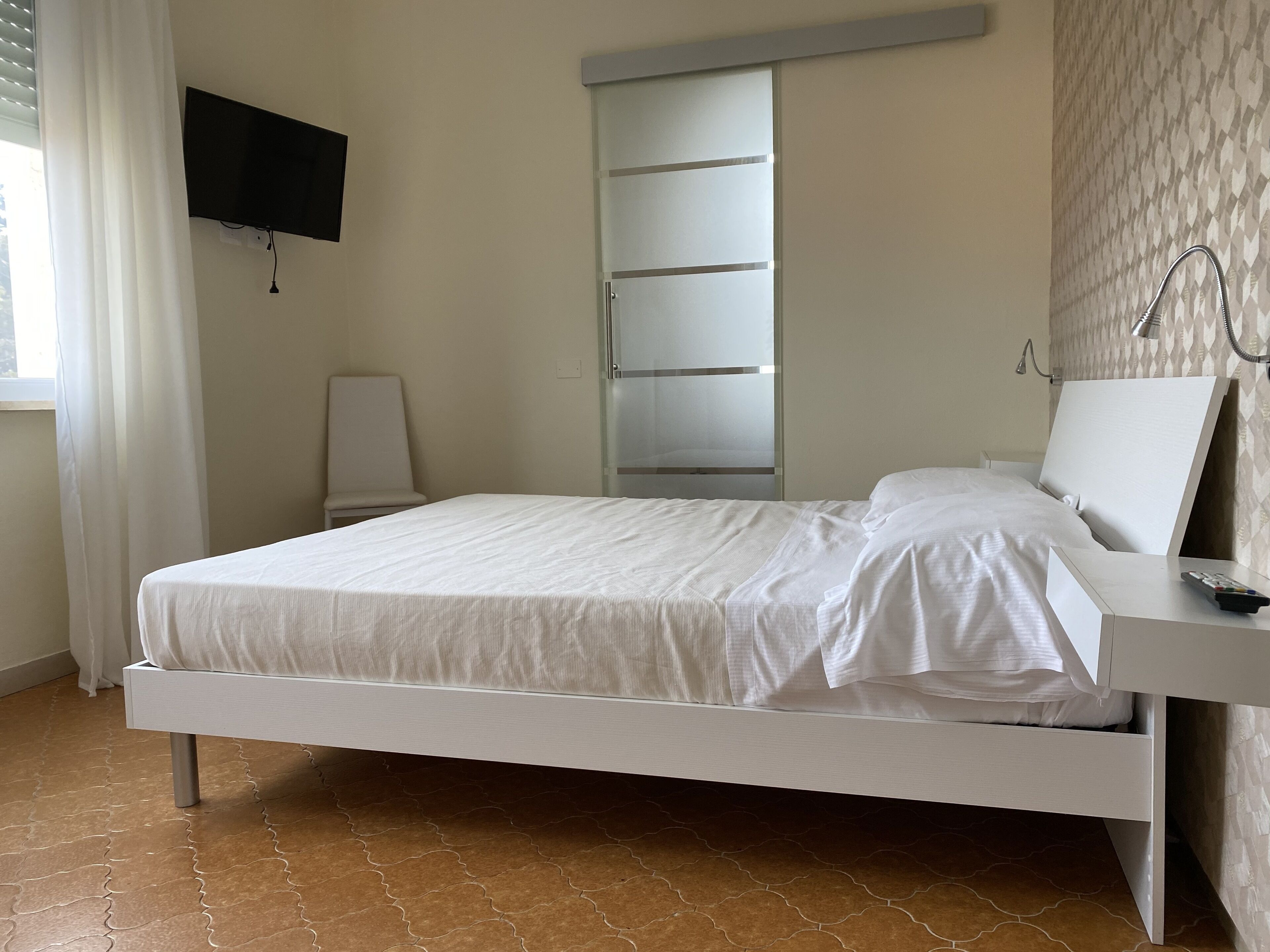 Una cama con colchón y almohadas blancas, una mesita de noche con un control remoto, un televisor montado en la pared y una puerta corredera para el armario.