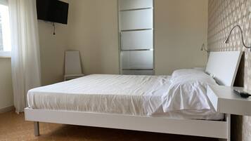 Una cama con colchón y almohadas blancas, una mesita de noche con un control remoto, un televisor montado en la pared y una puerta corredera para el armario.
