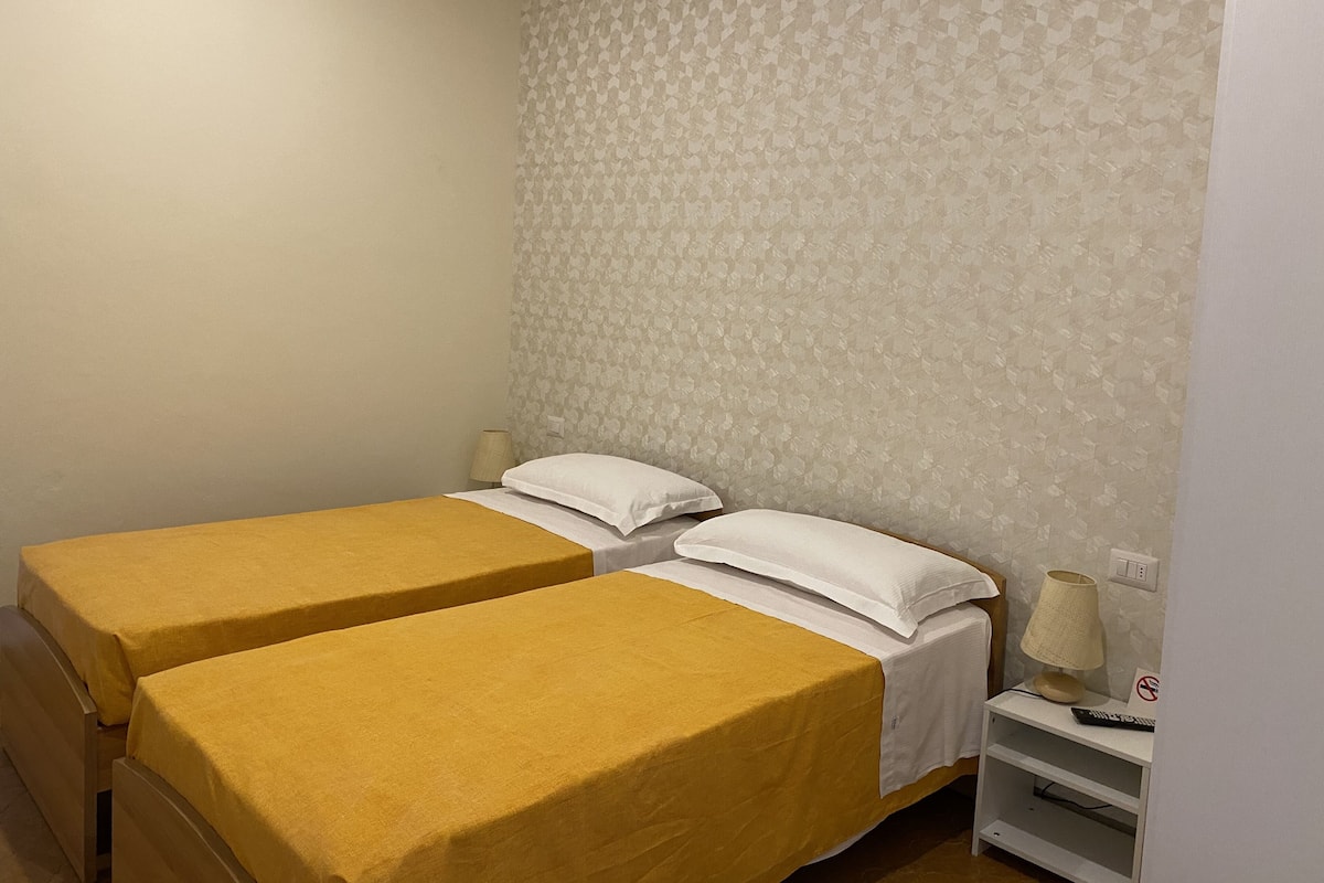 Ein Zimmer mit zwei Betten, jeweils mit gelber Bettdecke und weißen Kissen. Ein Nachttisch mit Lampe und einem Schild, das ein Rauchverbot anzeigt.