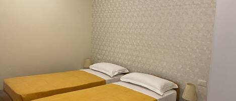 Une chambre avec deux lits, chacun recouvert d’une couvre-lit jaune et agrémenté d’oreillers blancs. On y trouve une table de chevet avec une lampe et un panneau indiquant qu’il est interdit de fumer.
