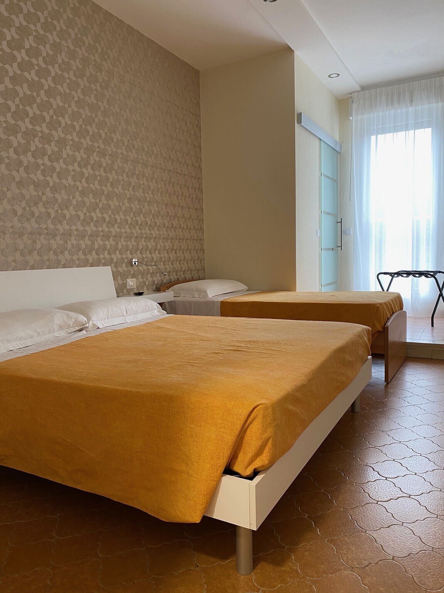 Quarto de hotel com duas camas, parede estampada, janela com cortinas e chão ladrilhado.