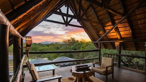 A wooden deck with a thatched roof, a table, two chairs, and a view of a swimming pool and greenery. - Vuyani Safari Lodge (Hoedspruit)