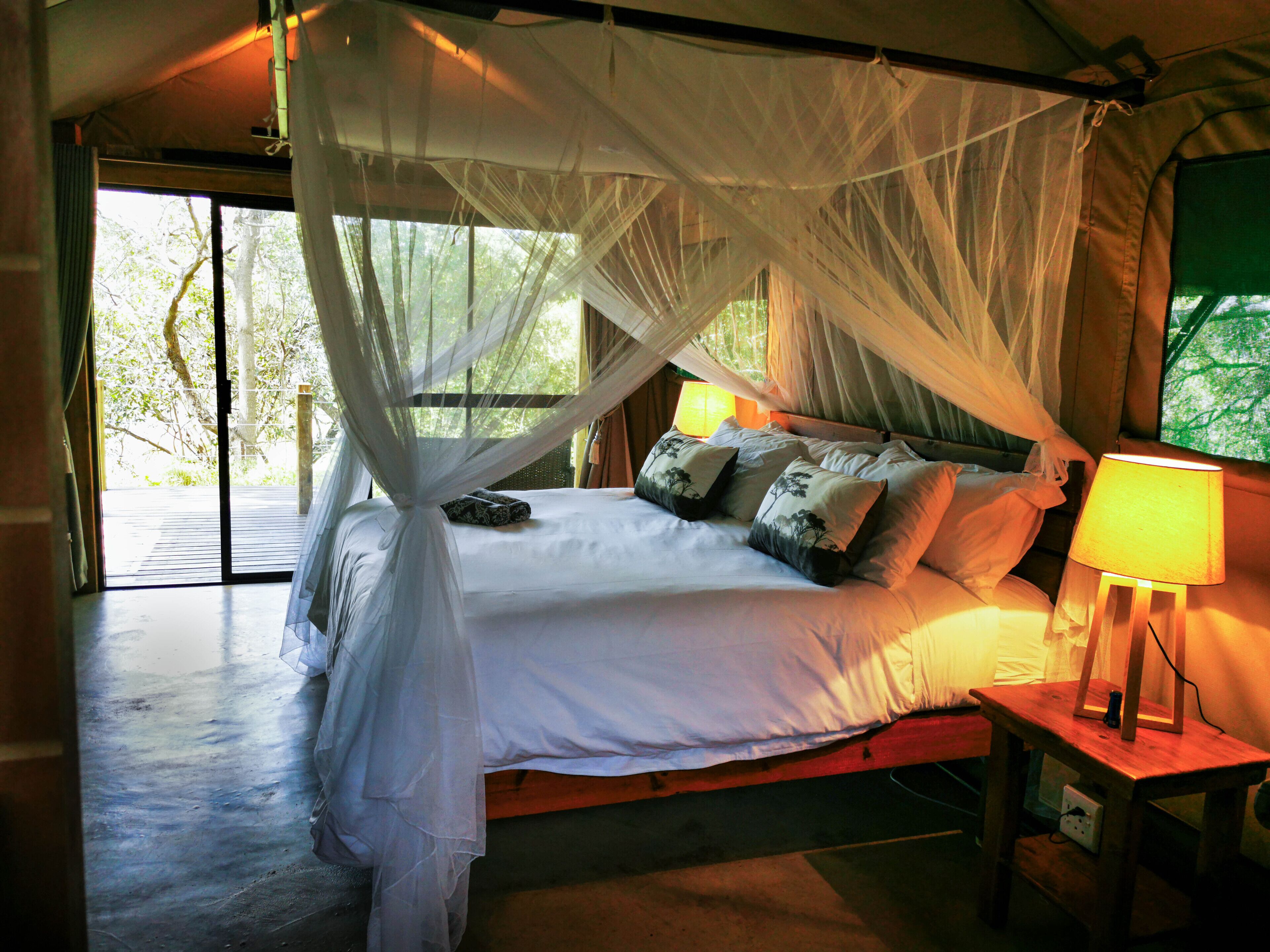 A bed with a canopy, pillows, a bedside table with a lamp, and a view of greenery outside.