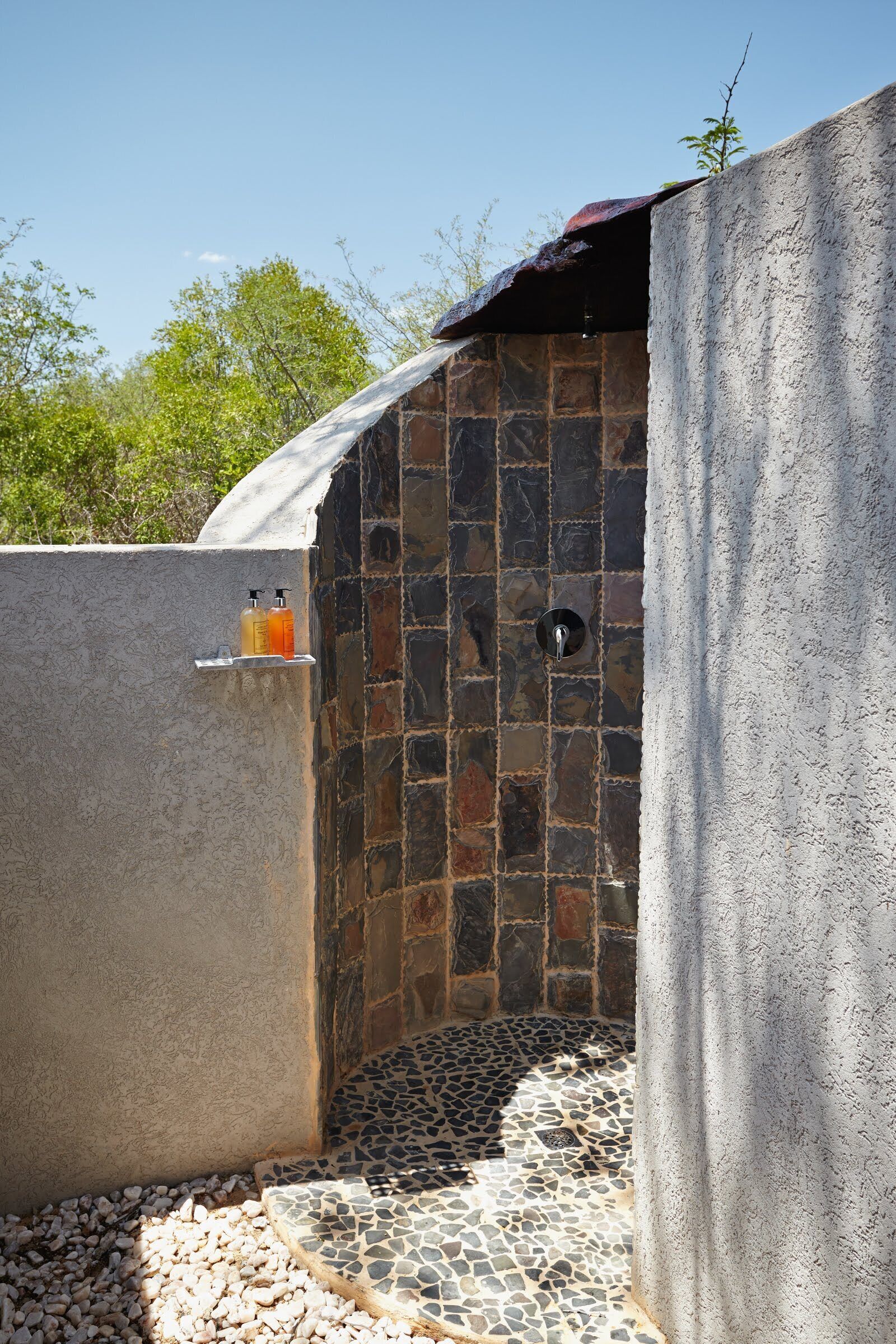A stone-walled outdoor shower area with a tiled floor, a small shelf with bottles, and a metal drain.