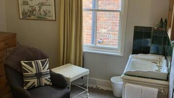 A small, cozy room with a single armchair, a small table, a window with curtains, a sink, and a mirror.