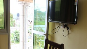 A room with a wooden desk, a chair, a television mounted on the wall, and a sliding glass door leading to a balcony. - Ndancy Hotel (Ebolowa)