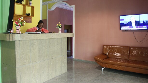 A reception area with a reception desk, a brown leather couch, a television mounted on the wall, and a decorative shelf with items. - Ndancy Hotel (Ebolowa)