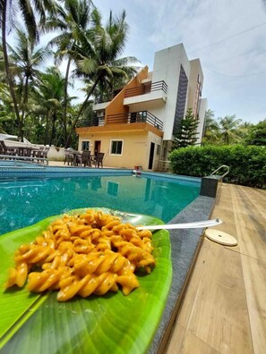 Vista de la piscina con un comedor, un edificio con balcones y palmeras al fondo.