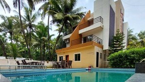 A poolside view with a dining table and chairs, a building with balconies, and palm trees in the background. - Royal Paradise Hotel (Alibag)