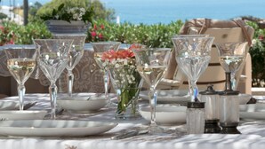 Ein Tisch ist gedeckt mit klarem Glas, weißen Tellern und einer Vase mit Blumen, im Hintergrund ist Meerblick.