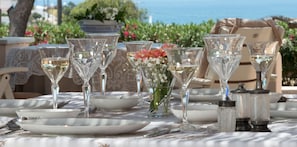 Una mesa dispuesta con vajilla de vidrio transparente, platos blancos y un jarrón con flores, con vista al mar.