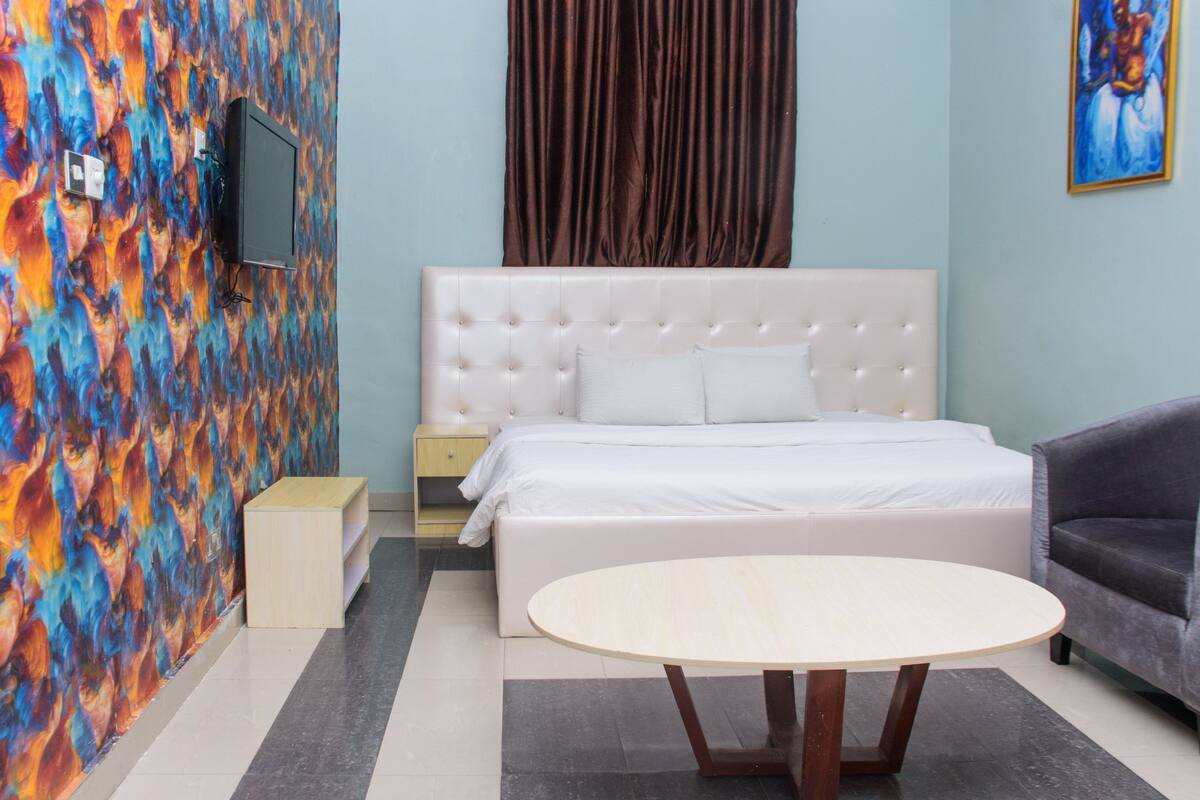 A bedroom with a white bed, a round wooden table, a grey sofa, a painting on the wall, and a TV mounted on the wall.