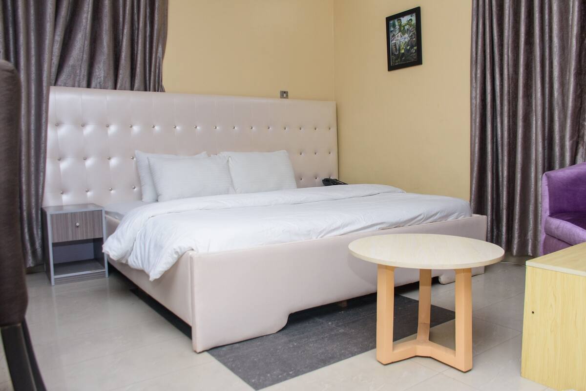 A hotel room with a large bed, a bedside table, a small round table, and a purple chair.