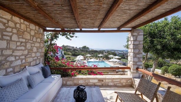 A stone-walled outdoor seating area with a wooden table, a black vase, and a view of a pool and landscape. - Sand Key Villa 1 - A beautiful cycladic residence 400m from the beach (Paros)