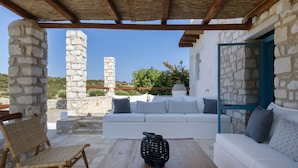 A stone-walled outdoor seating area with a wooden table, a woven chair, and a white sofa with patterned cushions. - Sand Key Villa 1 - A beautiful cycladic residence 400m from the beach (Paros)