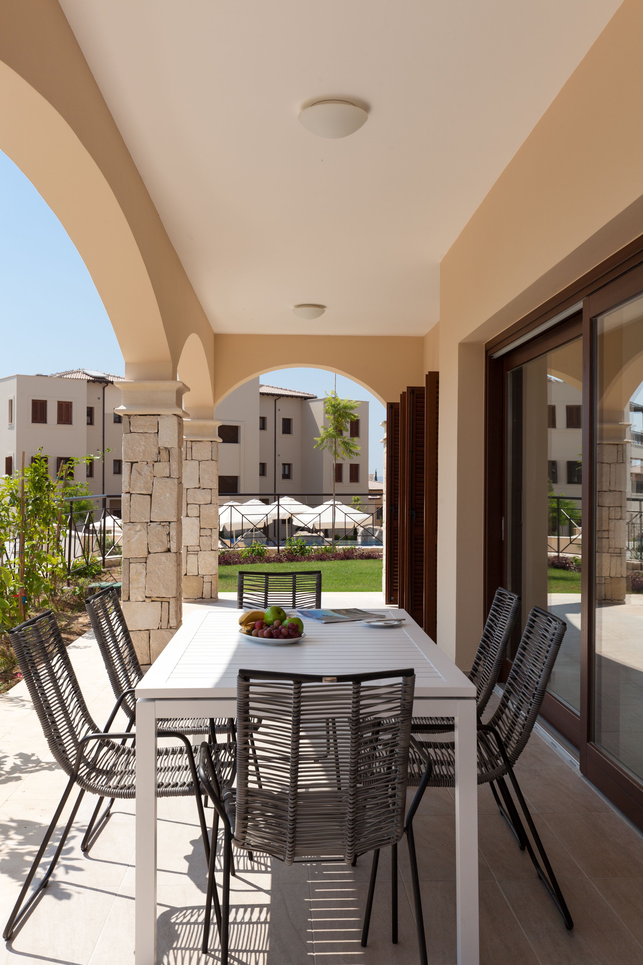 An outdoor patio with a white table and black wicker chairs, surrounded by stone pillars and a view of other buildings.