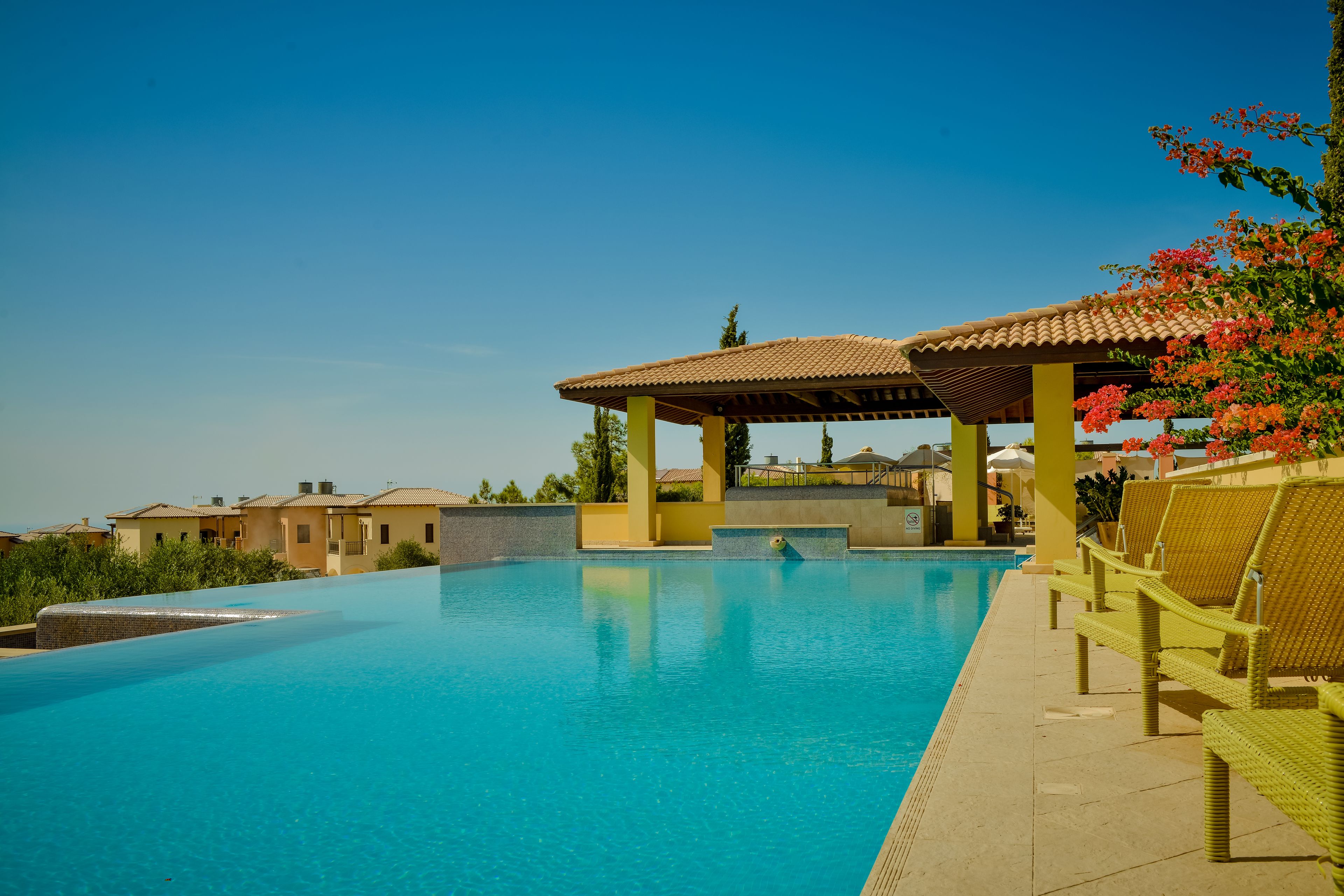 A clear blue swimming pool with a pergola and lounge chairs, surrounded by buildings.