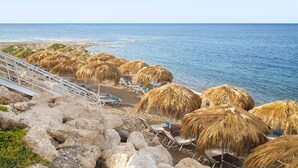 Una playa con sombrillas de paja, sillas de descanso, terreno rocoso y el océano.