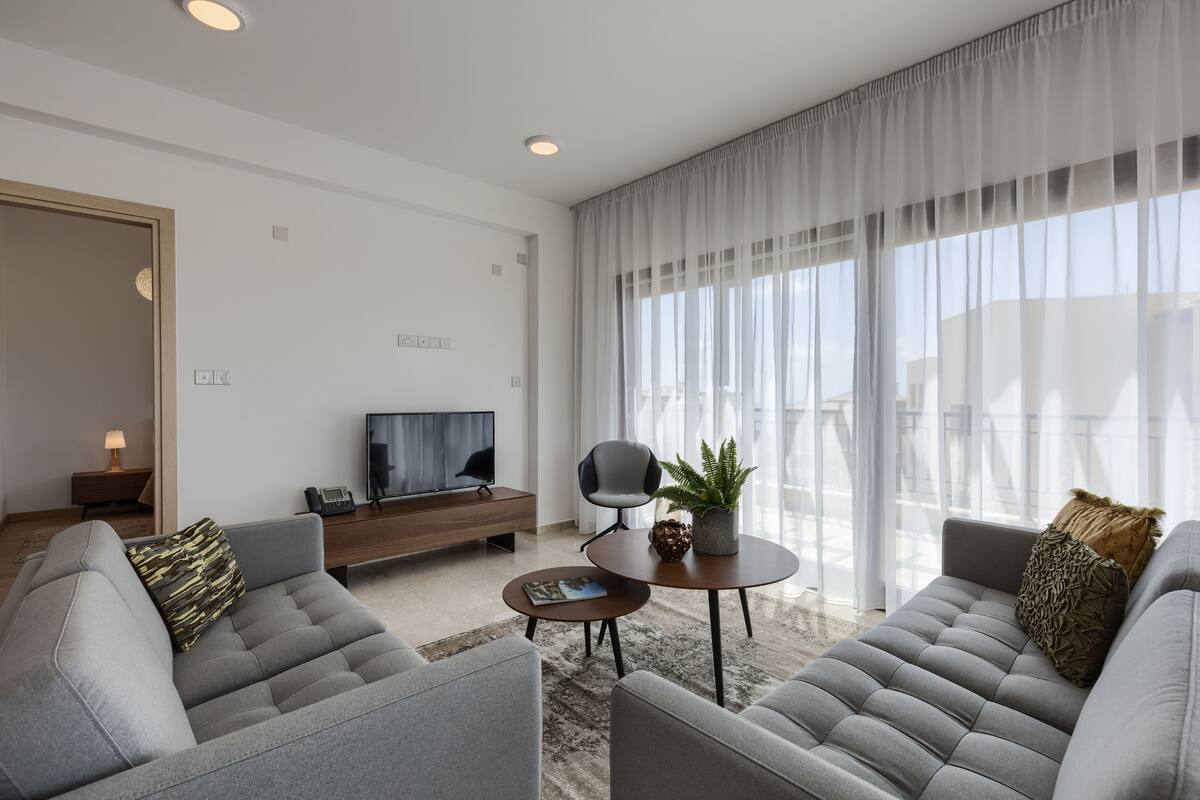 Ein modernes Wohnzimmer mit einer grauen Sofagarnitur, einem Couchtisch, einem Fernseher und einem großen Fenster mit transparenten Vorhängen.