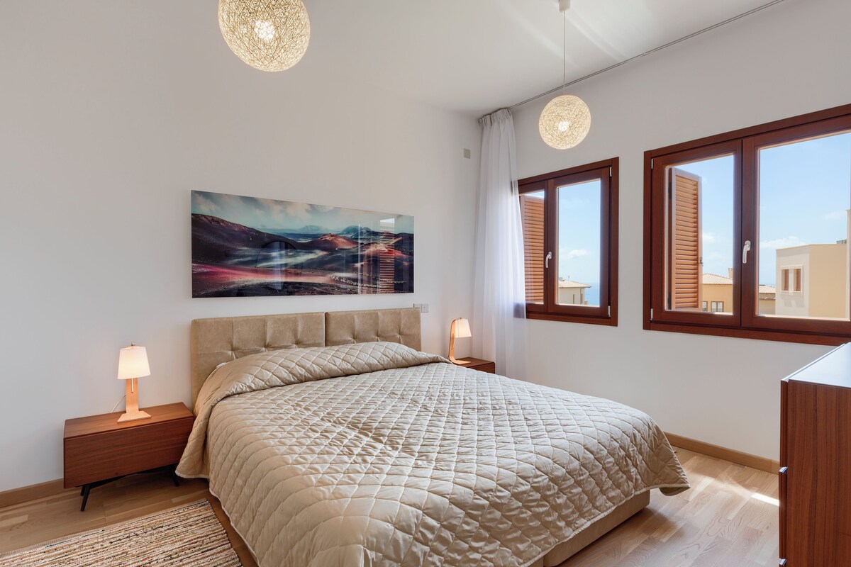 Un dormitorio moderno con una cama grande, dos mesitas de noche con lámparas, un cuadro grande en la pared y dos ventanas con marcos de madera.