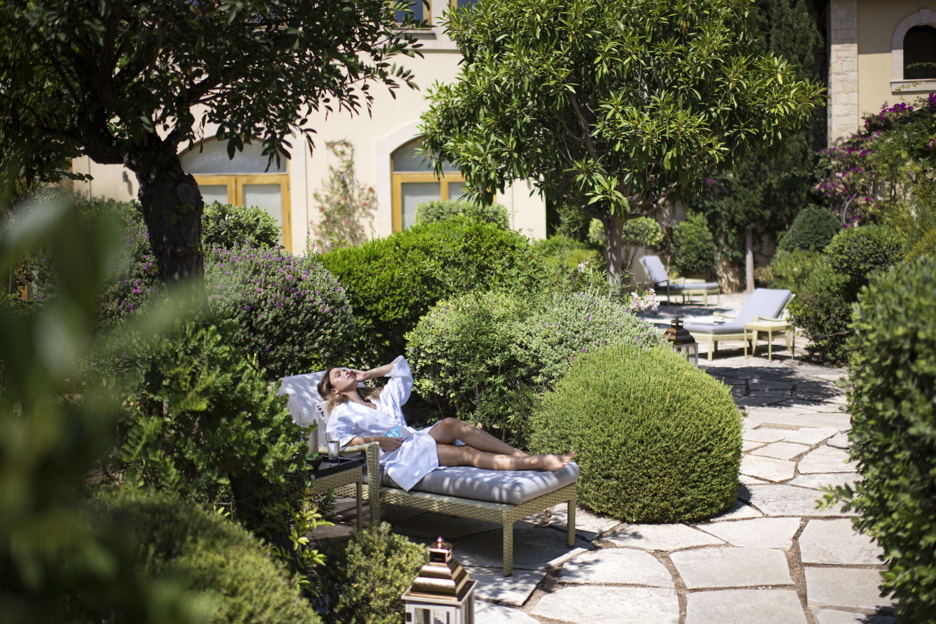 A person relaxing on a wicker lounge chair in a garden with stone pathways, surrounded by lush greenery and a building with a yellow door in the background.