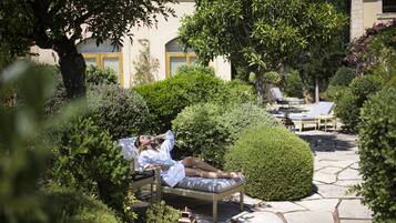 A person relaxing on a wicker lounge chair in a garden with stone pathways, surrounded by lush greenery and a building with a yellow door in the background.