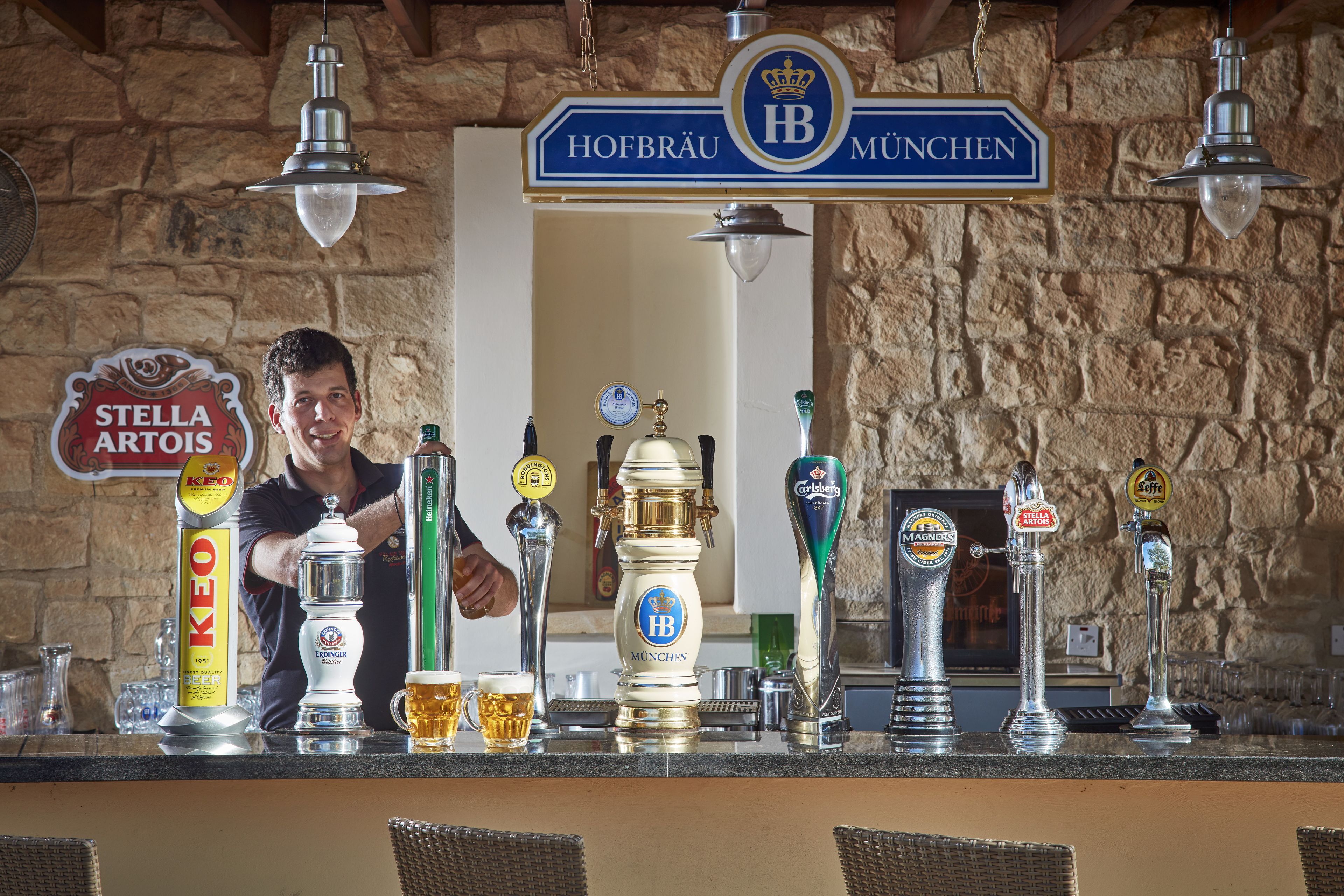 A bar with a stone wall background, a sign reading "Hofbräu München", and various beer taps including "Stella Artois" and "Grolsch".