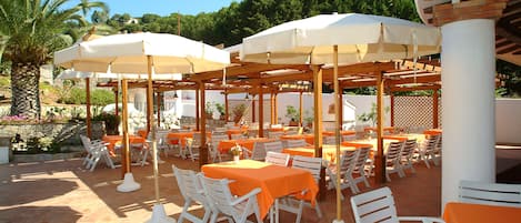 Un patio al aire libre con mesas y sillas, cubierto por sombrillas y rodeado de follaje.