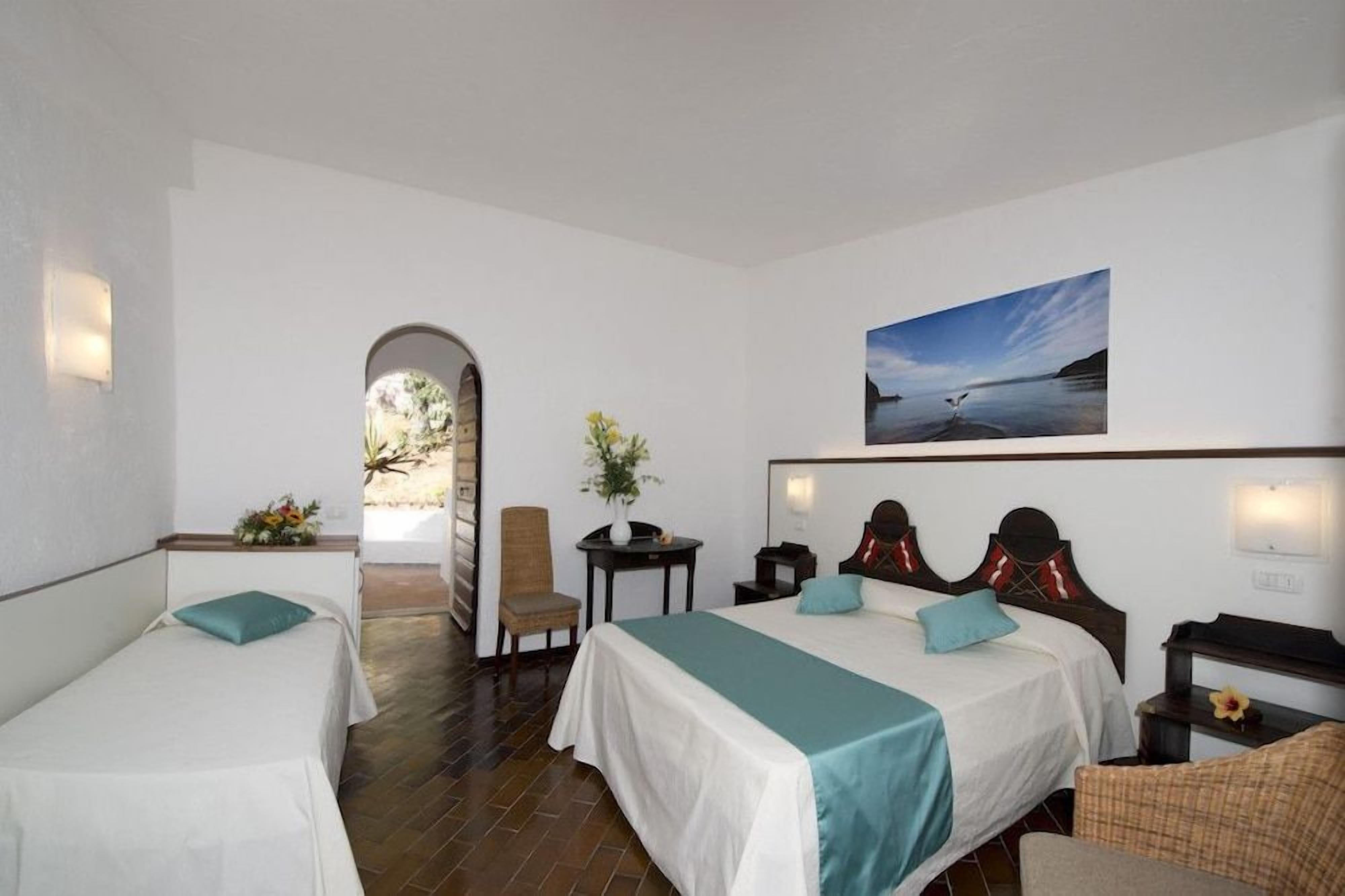 A hotel room with two beds, a desk, a chair, a small table, and a painting on the wall.