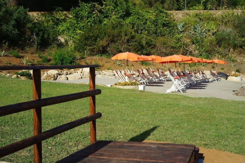 A poolside area with lounge chairs and orange umbrellas, surrounded by greenery and a stone wall.